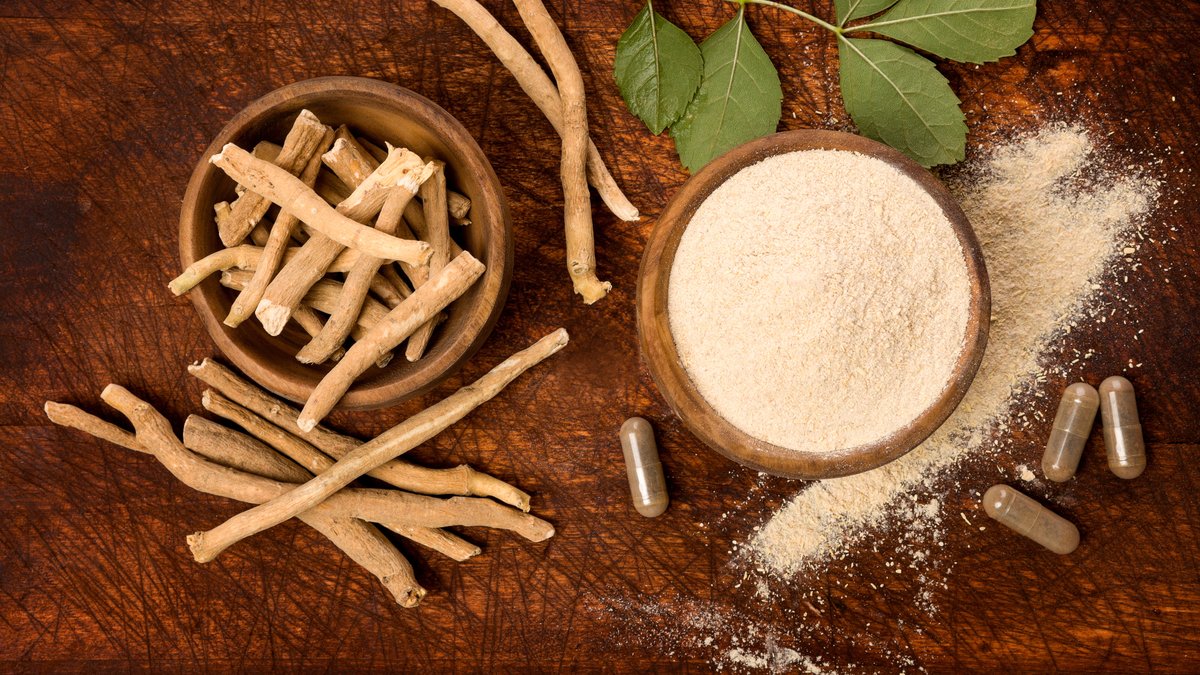 Auf einem Holztisch steht eine Schüssel mit Ashwagandha-Pulver, daneben liegen Ashwagandha-Wurzeln und Kapseln mit einem Ashwagandha-Präparat.