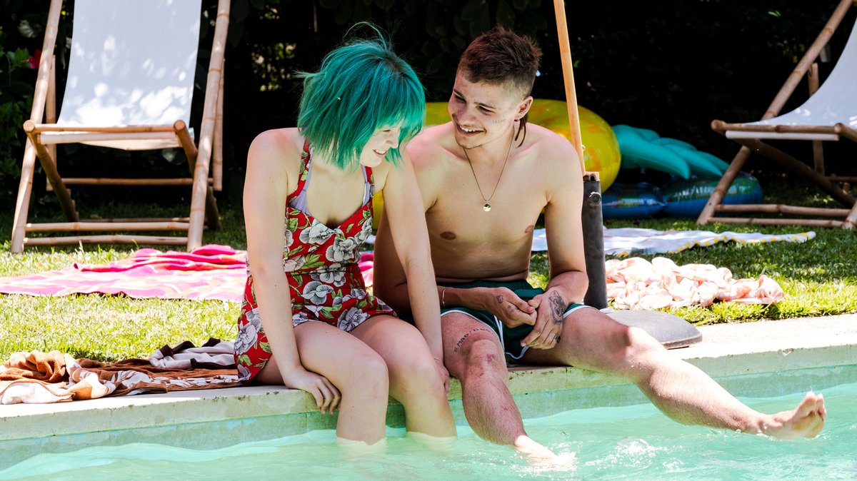Ein Mädchen mit grüner Perücke sitzt im Badedress neben einem jungen Mann an einem Swimmingpool: "Milla Meets Moses"-Filmszene