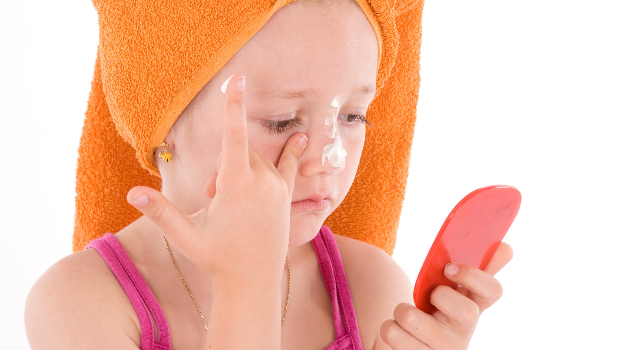 Ein Kind mit einem orangen Handtuchturban auf dem Kopf, schmiert sich mit einem Blick in den Handspiegel Creme ins Gesicht