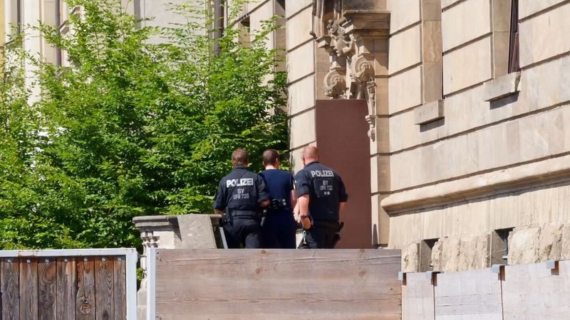 Archivbild 2. Juli 2025: Das Standbild aus einem Video zeigt, wie Polizeibeamte den Verdächtigen, der am Dienstag beim Stromversorger Überlandwerk Rhön in Mellrichstadt Kollegen mit einem Messer angegriffen, verletzt und eine tödlich verletzt haben soll, ins Gerichtsgebäude bringen. | Bild: picture alliance/dpa | Daniel Vogl Archivbild 2. Juli 2025: Das Standbild aus einem Video zeigt, wie Polizeibeamte den Verdächtigen, der am Dienstag beim Stromversorger Überlandwerk Rhön in Mellrichstadt Kollegen mit einem Messer angegriffen, verletzt und eine tödlich verletzt haben soll, ins Gerichtsgebäude bringen.