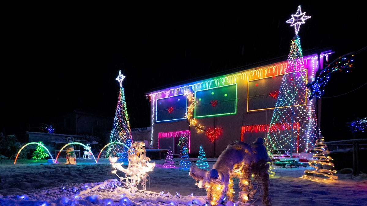 Ein mit bunter Weihnachtsbeleuchtung geschmücktes Haus, davor ein verschneiter Garten mit beleuchteter Weihnachtsdekoration