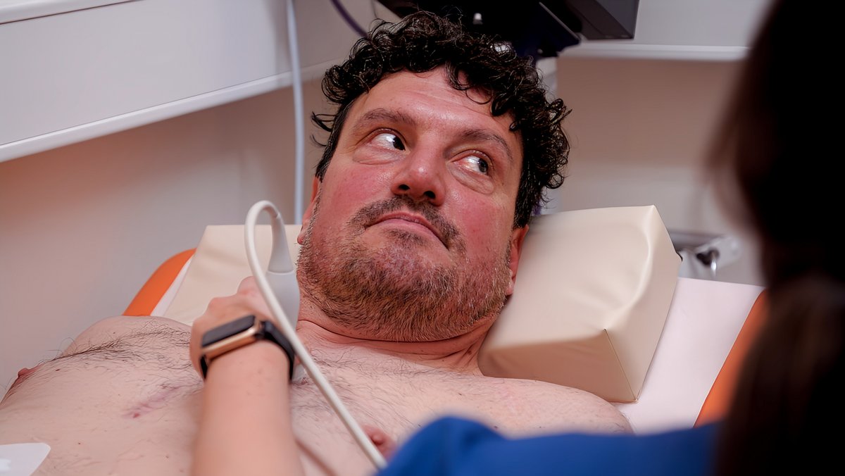 Mann liegt in einem Behandlungzimmer oberkörperfrei auf einer Liege, aus der Unschärfe fährt eine Person mit einem Ultraschallgerät über seine Brust.