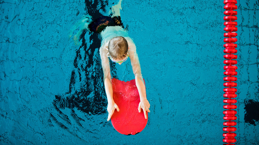 Ein Junge schwimmt in einem Schwimmbad, er hat ein rotes Schwimmbrett in der Hand.