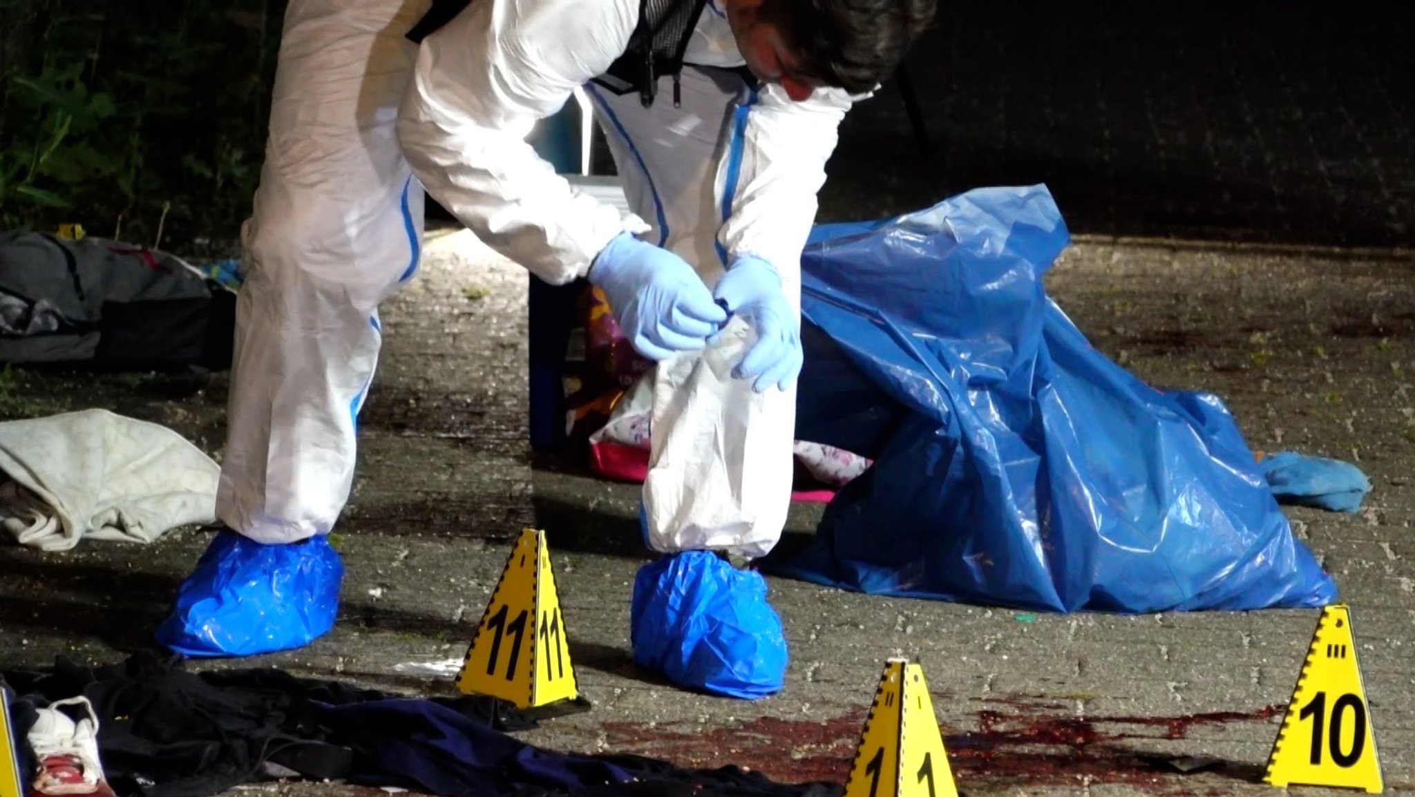 Ein Mann in einem weißen Schutzanzug sichert an einem Tatort Spuren. Ein blauer Plastiksack seht neben ihm. Gelbe Pylone mit Ziffern sind am Boden aufgestellt.  | Bild: BR Ein Mann in einem weißen Schutzanzug sichert an einem Tatort Spuren. Ein blauer Plastiksack seht neben ihm. Gelbe Pylone mit Ziffern sind am Boden aufgestellt.