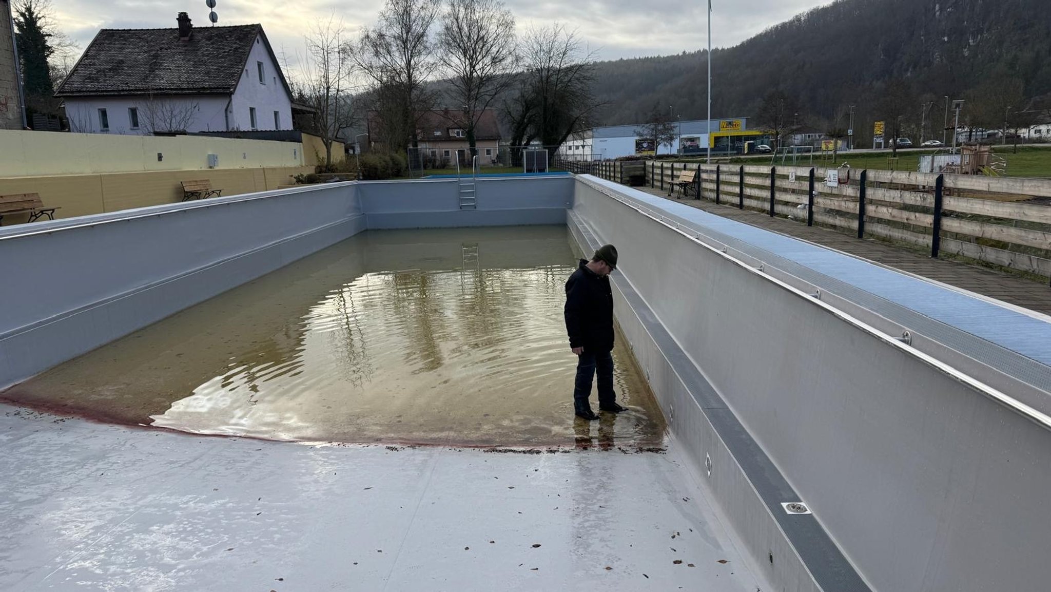 In einem fast leeren Schwimmbecken steht ein Mann und schaut auf eine Stufe an Beckenrand. Das restliche Wasser im Schwimmbecken ist voller Algen.