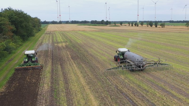 Vogelperspektive auf einen Acker im Emsland, auf dem die Wasserstoff-Protoyp-Traktoren von Fendt fahren. Beide haben einen weißen Kasten auf dem Dach, den Wasserstofftank. Im Hintergrund sind viele Windräder zu sehen. | Bild: BR Vogelperspektive auf einen Acker im Emsland, auf dem die Wasserstoff-Protoyp-Traktoren von Fendt fahren. Beide haben einen weißen Kasten auf dem Dach, den Wasserstofftank. Im Hintergrund sind viele Windräder zu sehen.