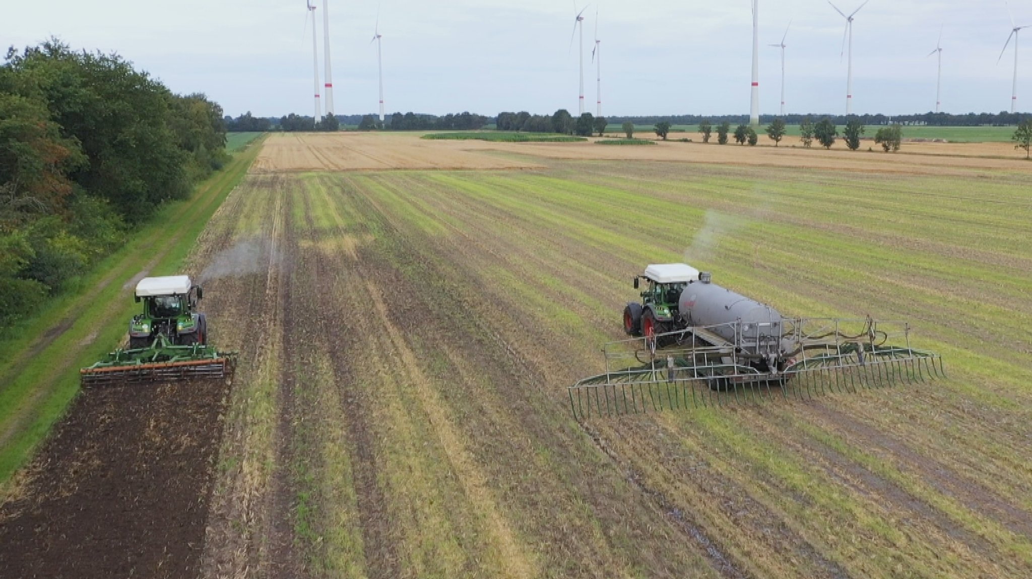 Vogelperspektive auf einen Acker im Emsland, auf dem die Wasserstoff-Protoyp-Traktoren von Fendt fahren. Beide haben einen weißen Kasten auf dem Dach, den Wasserstofftank. Im Hintergrund sind viele Windräder zu sehen.