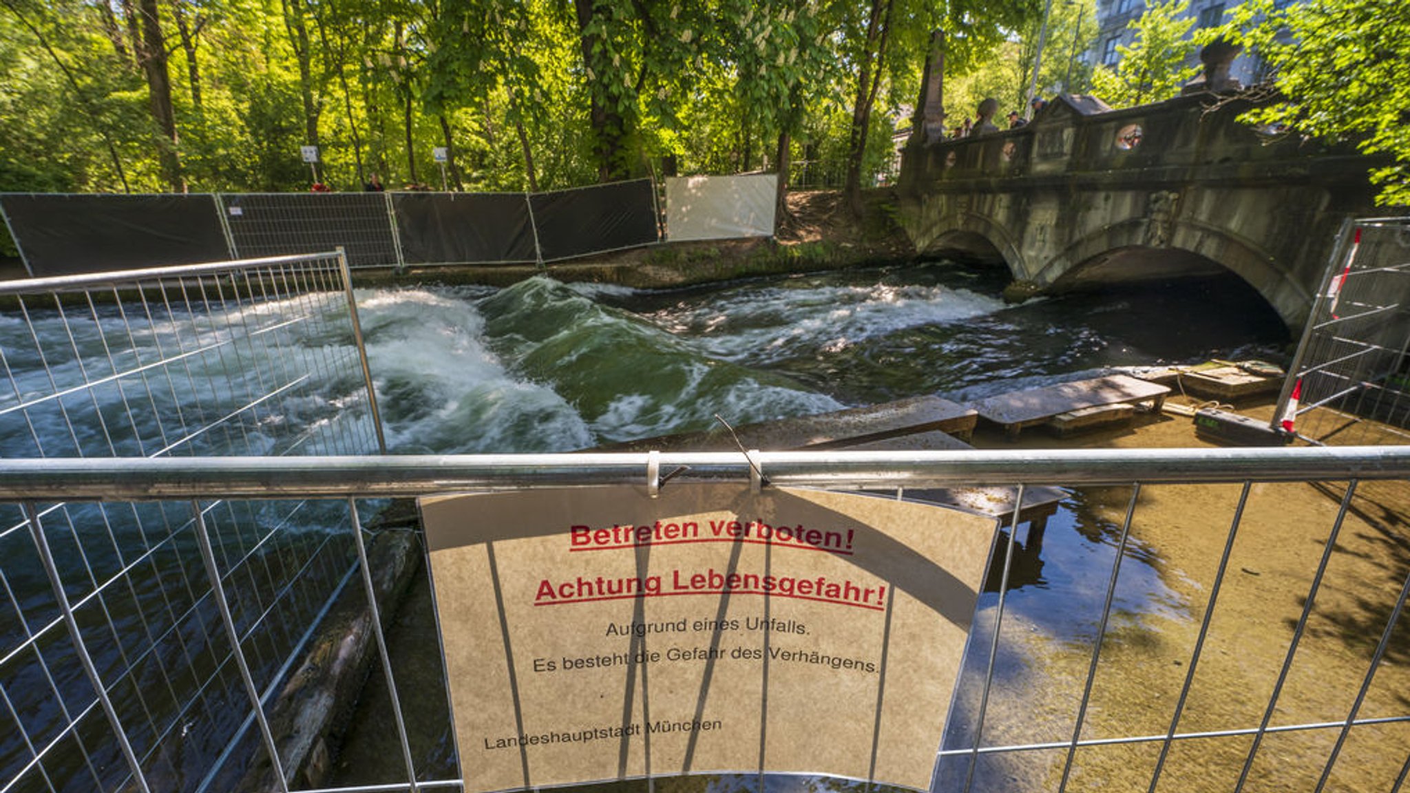 ARCHIV - 29.04.2025, Bayern, München: Ein Schild mit der Aufschrift ·Betreten verboten! Achtung Lebensgefahr! - Aufgrund eines Unfalls. - Es besteht die Gefahr des Verhängens.· hängt an einem Absperrgittter am Ufer unweit der künstliche Eisbachwelle im Englischen Garten. Eine Surferin, die vor Kurzem auf der Eisbachwelle im Englischen Garten stürzte, war im Krankenhaus verstorben. (zu dpa: «Surfer fordern Freigabe der Eisbachwelle») Foto: Peter Kneffel/dpa +++ dpa-Bildfunk +++