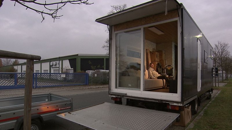 Ein Mann sitzt auf einer Couch. Diese befindet sich im Inneren eines Lkw-Anhängers, der wie ein Wohnzimmer eingerichtet ist. Der Anhänger steht in einem Industriegebiet. | Bild: BR/Carlo Schindhelm Ein Mann sitzt auf einer Couch. Diese befindet sich im Inneren eines Lkw-Anhängers, der wie ein Wohnzimmer eingerichtet ist. Der Anhänger steht in einem Industriegebiet.