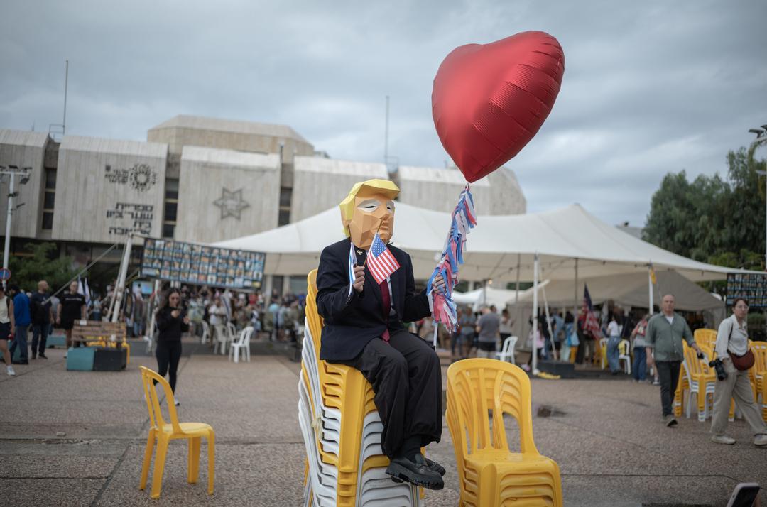 09.10.2025, Israel, Tel Aviv: Eine Person trägt eine Maske, die US-Präsident Trump ähnelt, und hält die US-Flagge und einen herzförmigen Ballon in der Hand, während sich Menschen auf dem Geiselplatz versammeln, um das Waffenstillstandsabkommen zu feiern. 