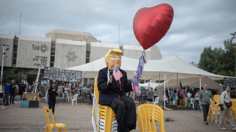 09.10.2025, Israel, Tel Aviv: Eine Person trägt eine Maske, die US-Präsident Trump ähnelt, und hält die US-Flagge und einen herzförmigen Ballon in der Hand, während sich Menschen auf dem Geiselplatz versammeln, um das Waffenstillstandsabkommen zu feiern. | Bild: dpa-Bildfunk/Ilia Yefimovich 09.10.2025, Israel, Tel Aviv: Eine Person trägt eine Maske, die US-Präsident Trump ähnelt, und hält die US-Flagge und einen herzförmigen Ballon in der Hand, während sich Menschen auf dem Geiselplatz versammeln, um das Waffenstillstandsabkommen zu feiern.