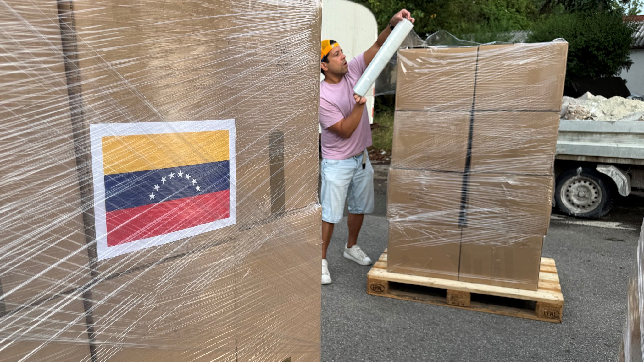 Ein Mann sichert einen Stapel gepackter Pakete mit durchsichtiger Folie. Auf einem der Pakete klebt eine venezolanische Flagge. | Bild: BR / David Herting Ein Mann sichert einen Stapel gepackter Pakete mit durchsichtiger Folie. Auf einem der Pakete klebt eine venezolanische Flagge.