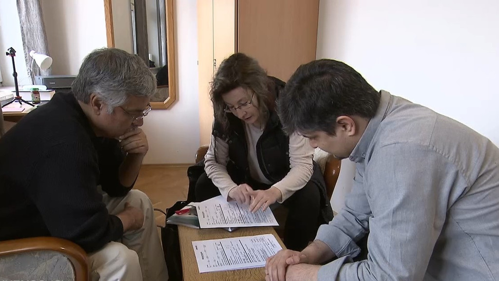 Ein Frau hilft zwei Männern beim Ausfüllen von Formularen, die auf einem Tisch liegen.