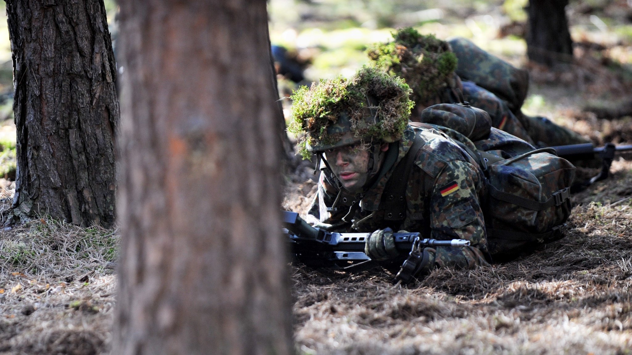 Soldaten in Tarnklamotten liegen zwischen Bäumen auf dem Waldboden. In ihren Händen halten sie Gewehre. Auf ihren Helme haben sie Pflanzen angebracht. (Symbolbild) | Bild: picture alliance / dpa | Maurizio Gambarini Soldaten in Tarnklamotten liegen zwischen Bäumen auf dem Waldboden. In ihren Händen halten sie Gewehre. Auf ihren Helme haben sie Pflanzen angebracht. (Symbolbild)