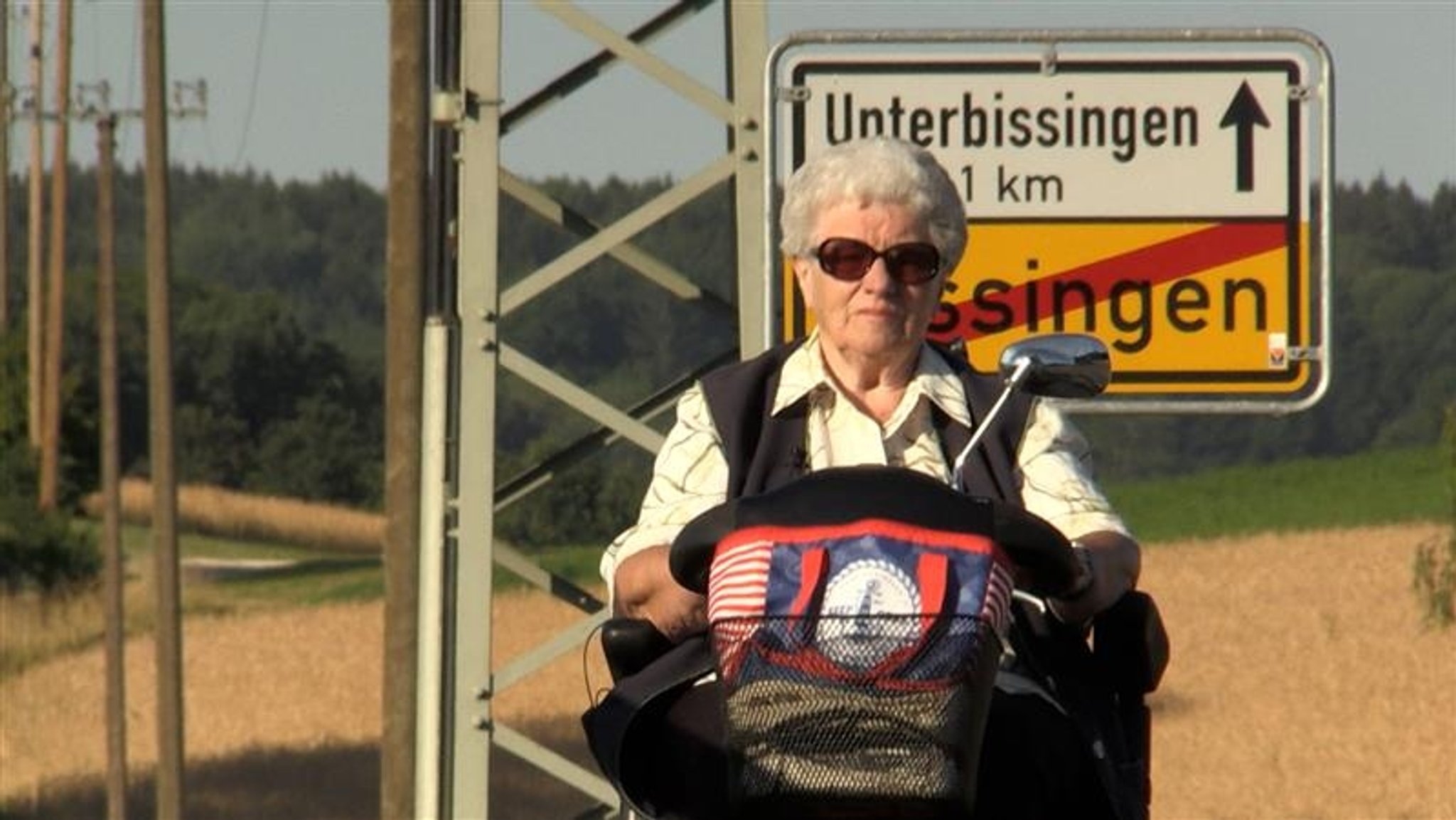 Anni Kalchgruber auf dem Weg zum Rathaus in Bissingen. Im Hintergrund das Ortsschild von Bissingen, Felder und ein Wald