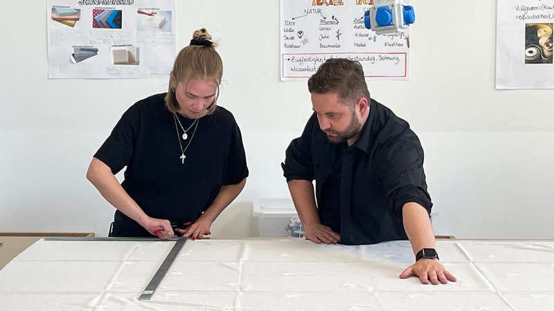 Ein Ausbilder und eine Auszubildende schneiden einen Stoff zu. | Bild: BR / Julia Demel Ein Ausbilder und eine Auszubildende schneiden einen Stoff zu.