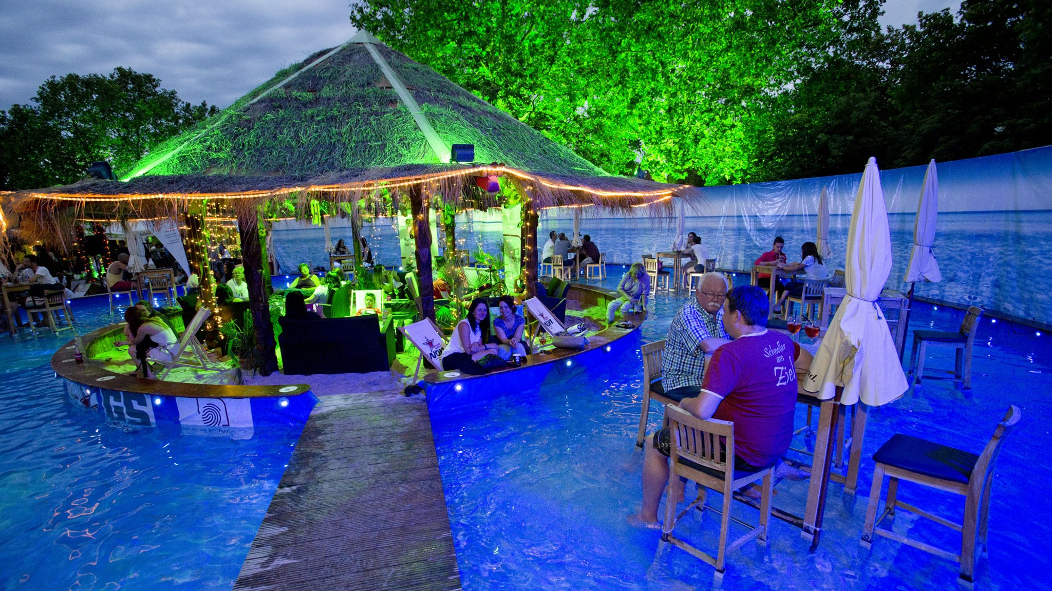 Eine Strandbar sowie Bartische und -stühle in einem Pool in der Abenddämmerung.