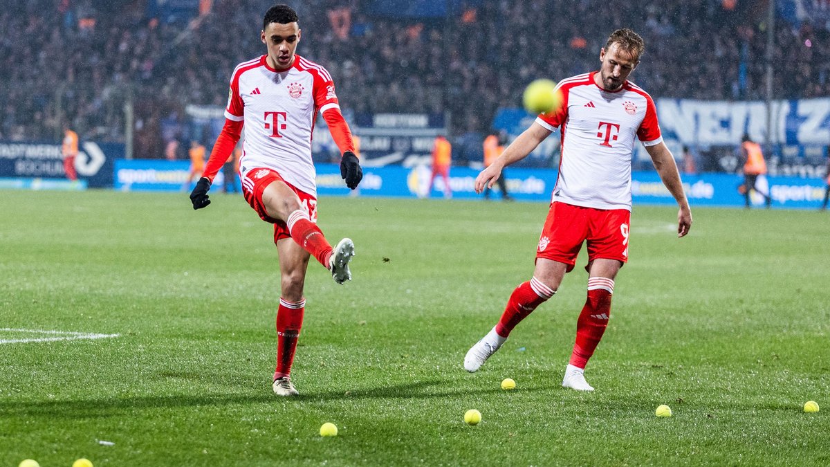 Jamal Musiala und Harry Kane vom FC Bayern räumen Tennisbälle vom Spielfeld.
