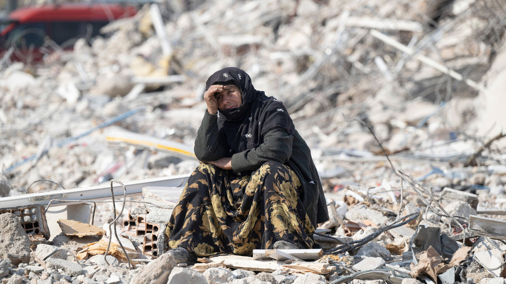 Eine Person sitzt zwischen den Trümmern und beobachtet die Bergung von Opfern. 