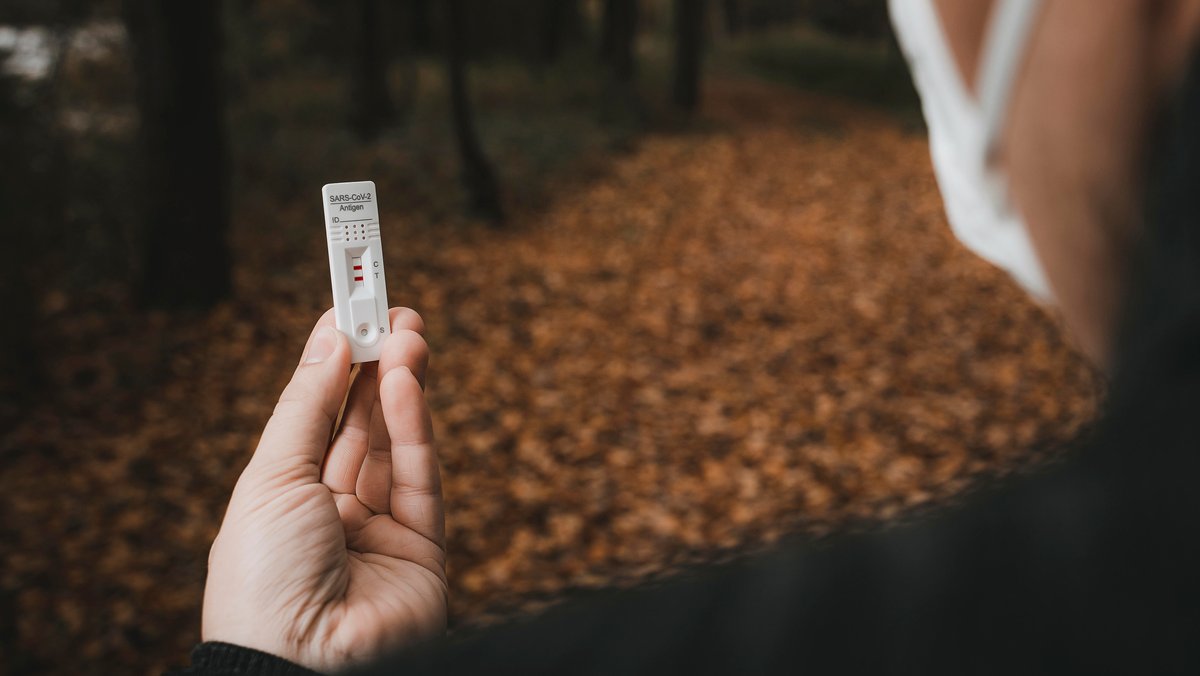 Ein Mann hält in einem herbstlichen Wald einen Covid-19-Schnelltest mit Testergebnis "positiv" in der Hand (Symbolbild)