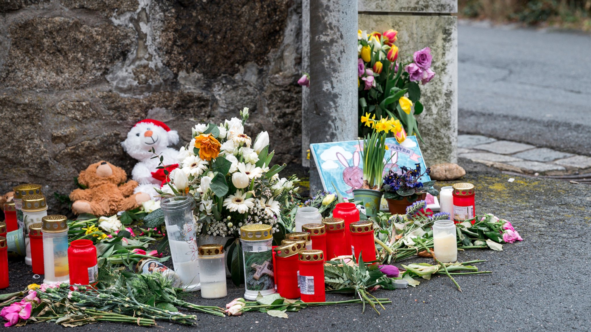 Blumen, Grabkerzen, ein gemaltes Bild und Kuscheltiere sind auf der Straße bei einer Mauer abgelegt. 