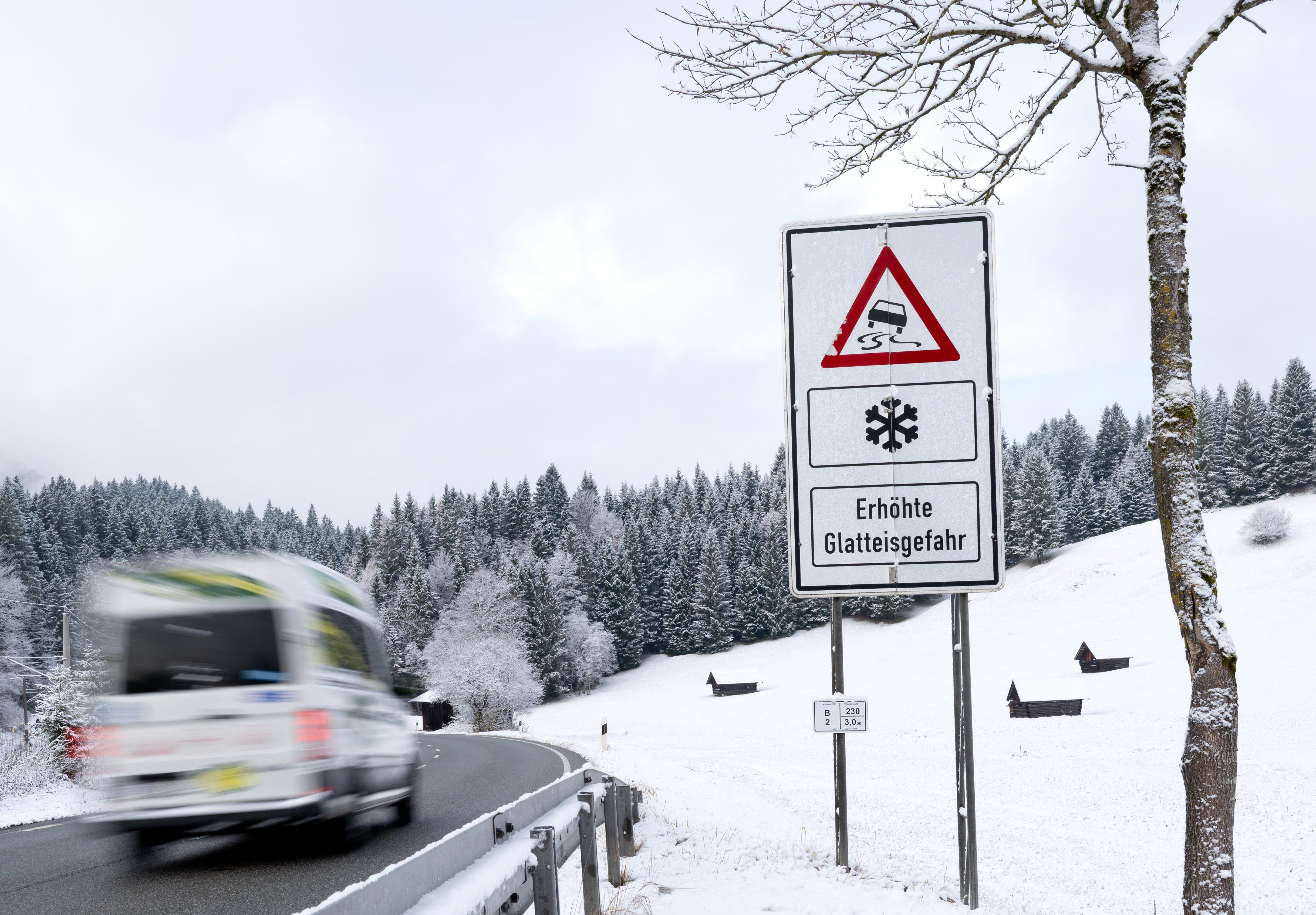 18.11.2025, Bayern, Garmisch-Partenkirchen: Ein Lieferwagen fährt über eine Straße an einem Schild mit der Aufschrift „Erhöhte Glatteisgefahr“ entlang.