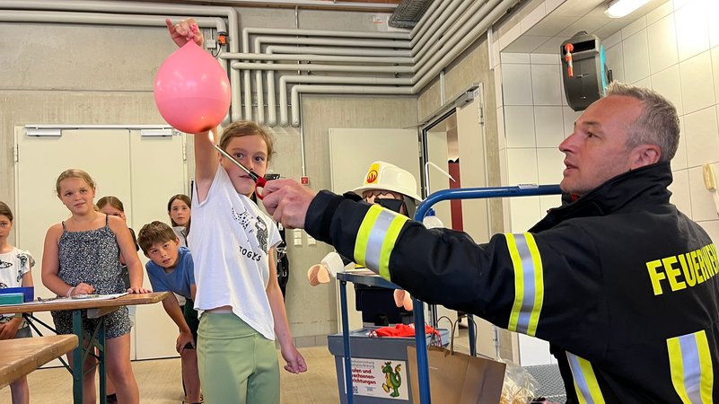 Ein Mädchen hält einen rosa Luftballon in der Hand. Ein Feuerwehrmann in Uniform hält eine Flamme darunter.