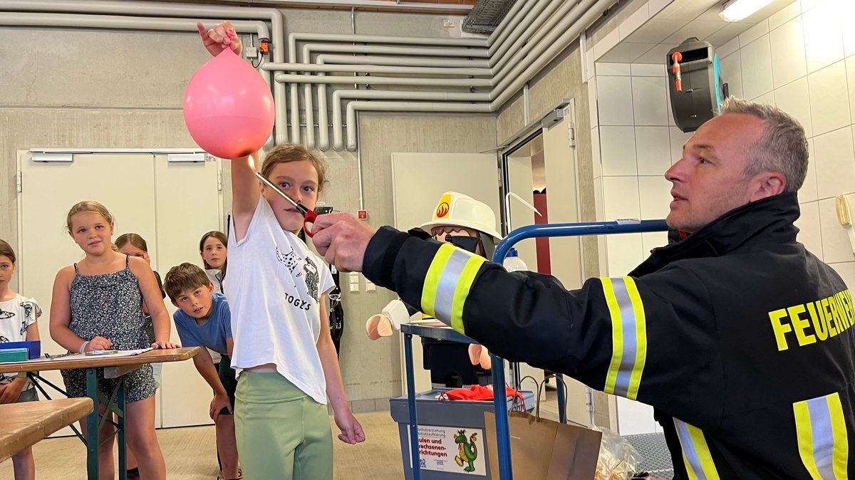 Ein Mädchen hält einen rosa Luftballon in der Hand. Ein Feuerwehrmann in Uniform hält eine Flamme darunter.