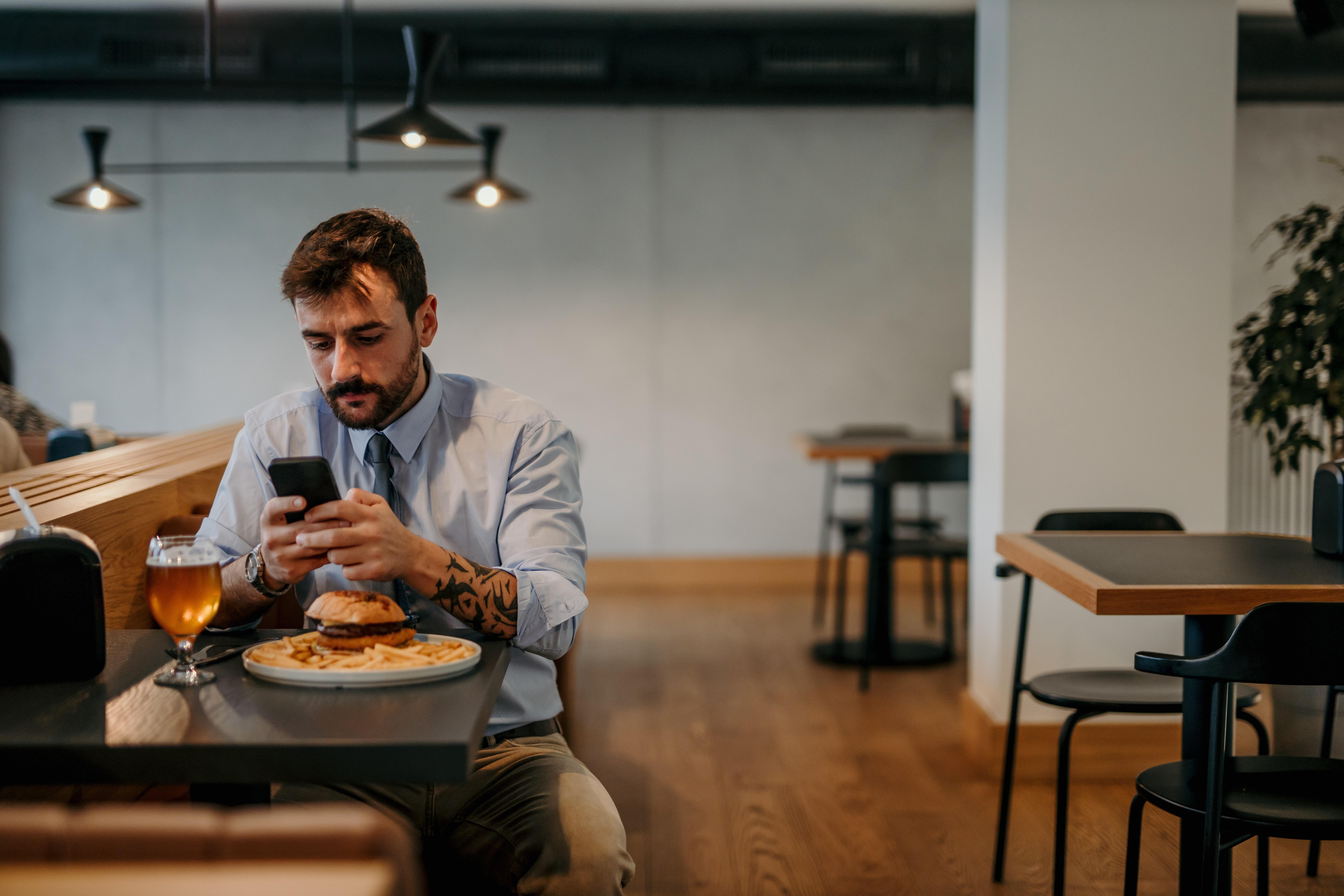 Ein Mann sitzt in einem Restaurant bei einem Burger und einem Bier, hält ein Smartphone in der Hand und tippt darauf eine Nachricht.
