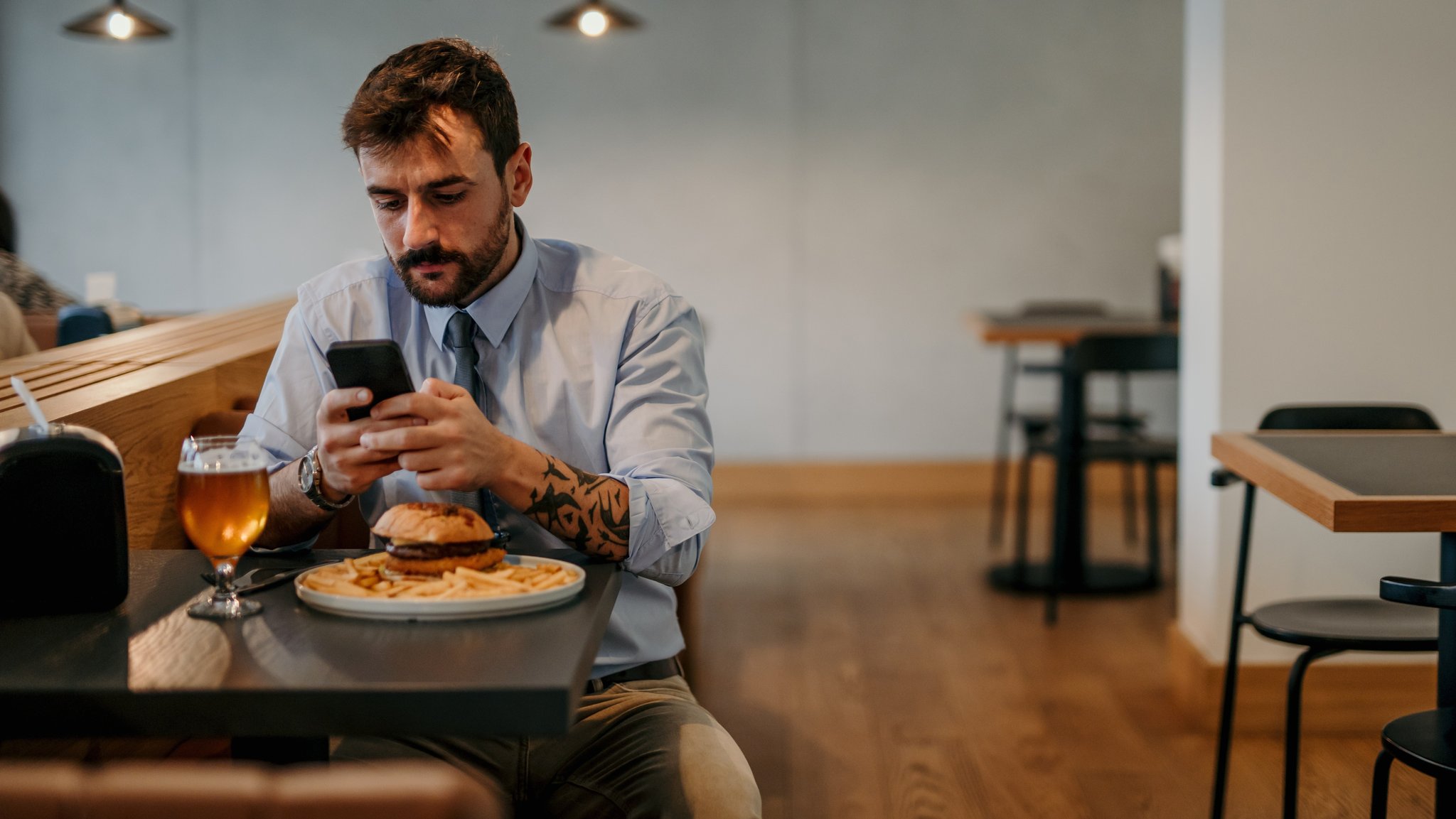 Ein Mann sitzt in einem Restaurant bei einem Burger und einem Bier, hält ein Smartphone in der Hand und tippt darauf eine Nachricht.