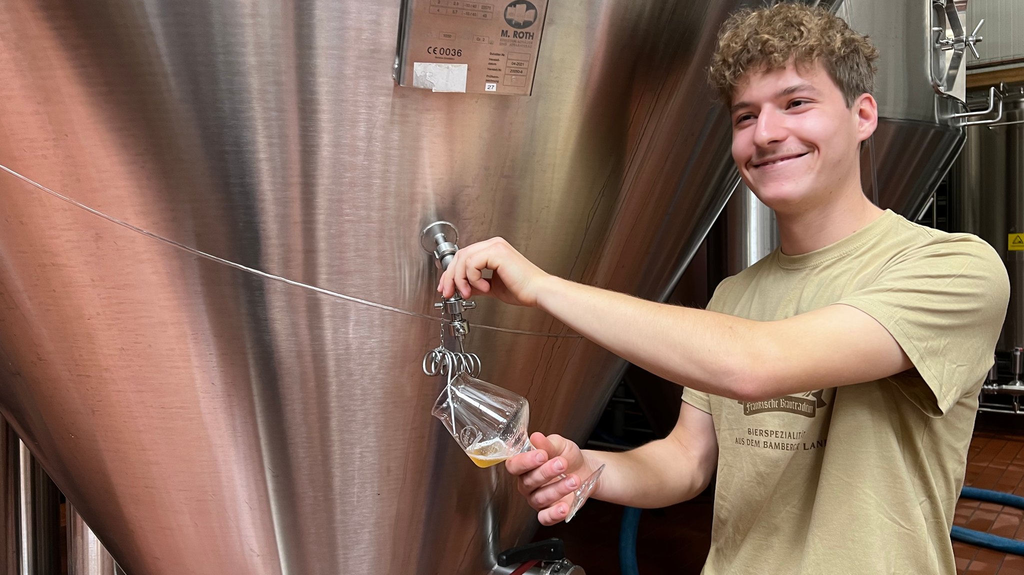 Ein junger Mann steht an einem Biertank und lächelt. Er hält mit einer Hand ein Pilsglas in der Hand. Mit der anderen Hand betätigt er den Hahn des Tanks. Es fließt Bier aus dem Tank in das Glas.