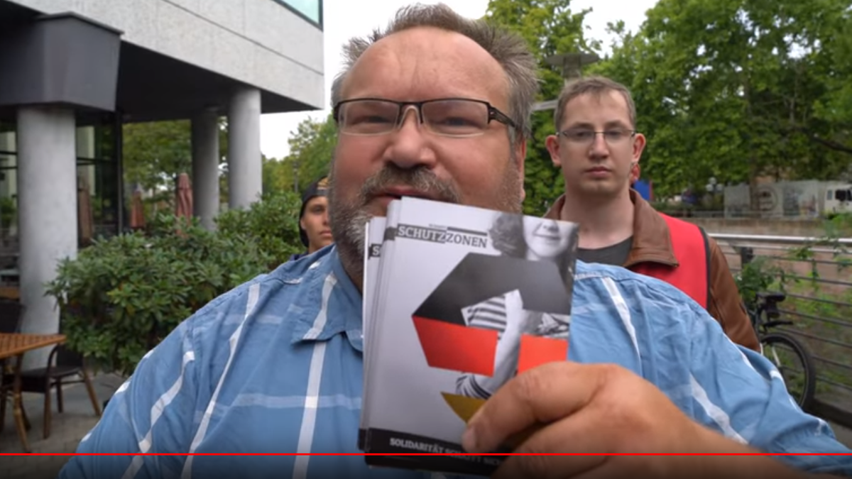 Ein korpulenter Mann hält Flyer in die Kamera, auf denen ist ein Logo in schwarz-rot-gold zu sehen. Das Wort "Schutzzone" ist lesbar.