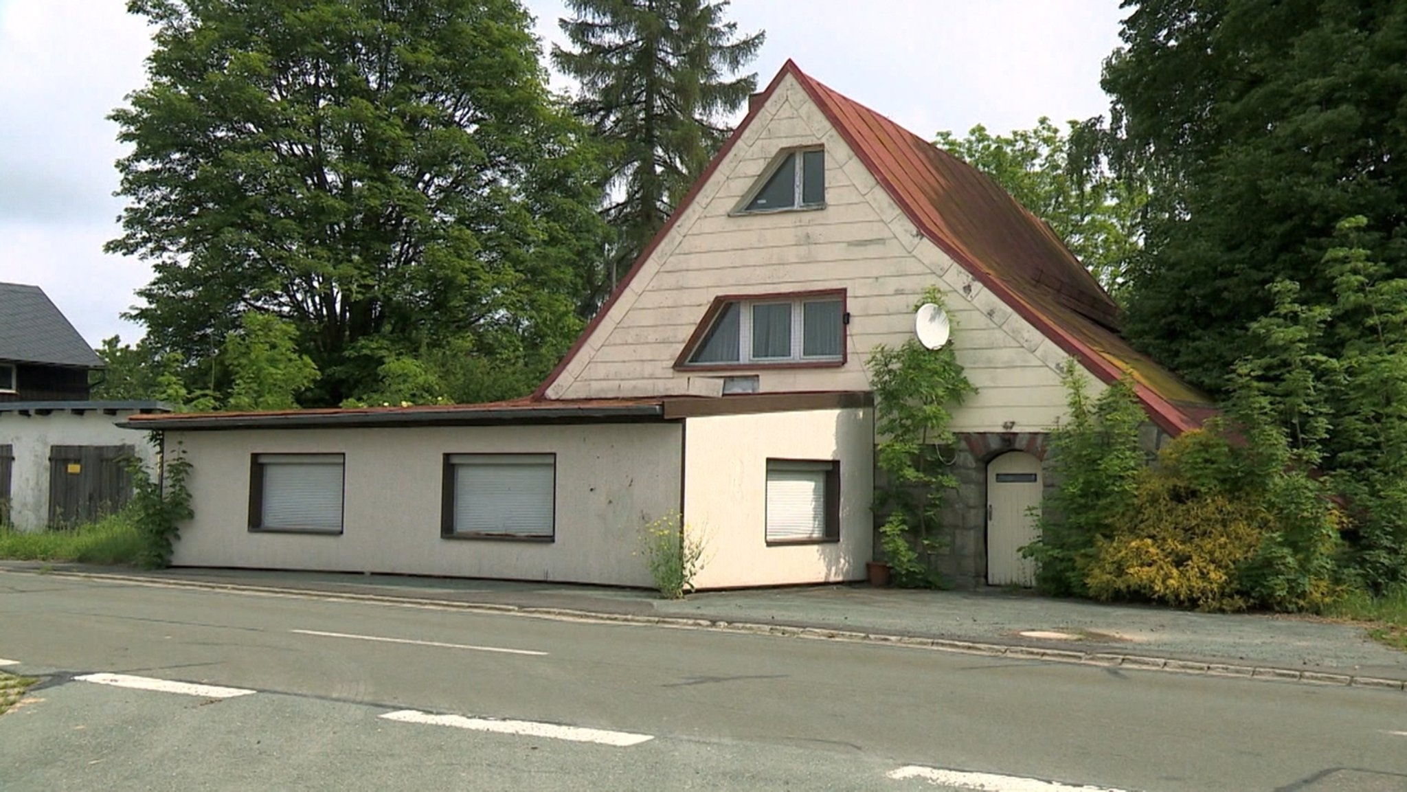 Ein Haus mit rotem Dach, davor ein flacher Anbau, dahinter hohe Laubbäume. 