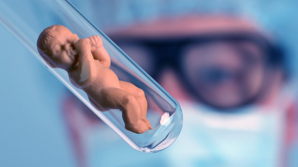 Symbolbild genverändertes Baby: Babypüppchen in einem Reagenzglas, Mediziner im Hintergund,