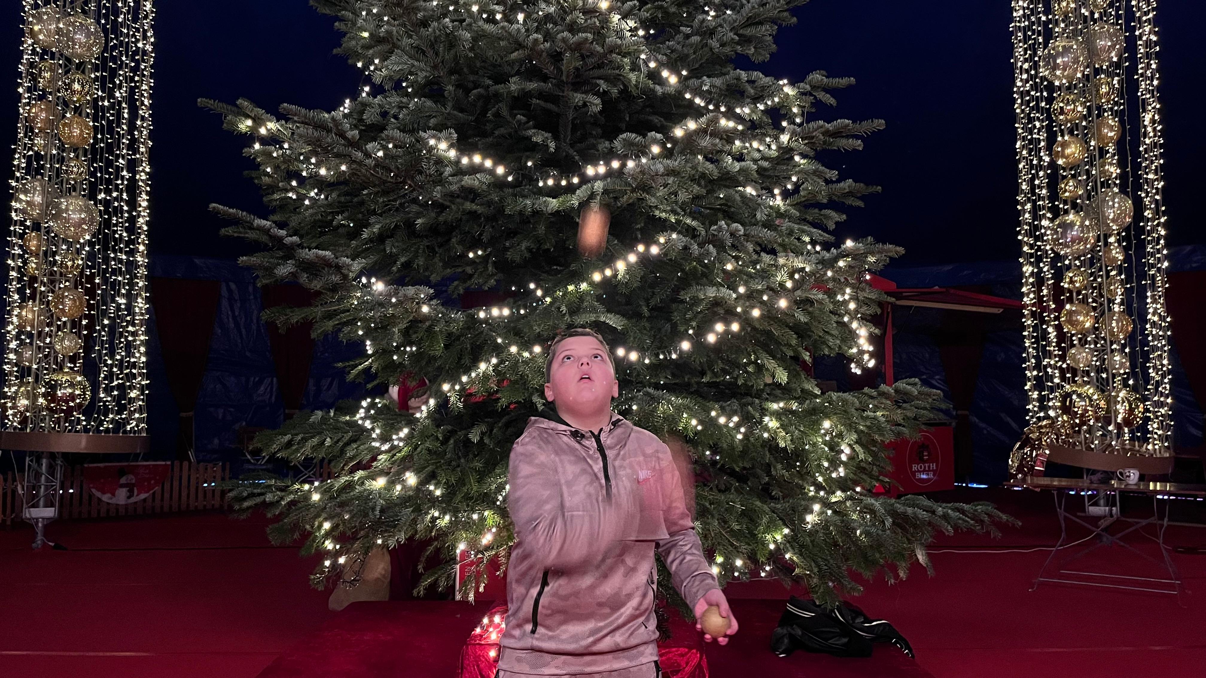 Ein Junge jongliert mit drei Bällen vor einem Weihnachtsbaum mit erleuchteter Lichterkette
