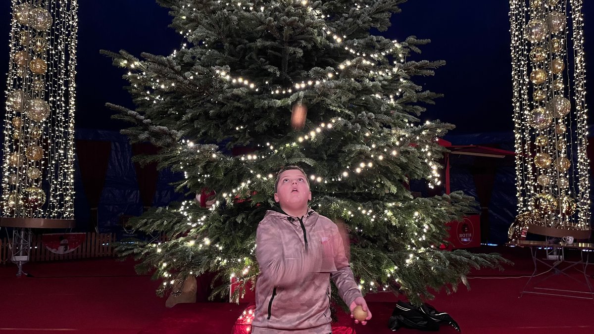 Ein Junge jongliert mit drei Bällen vor einem Weihnachtsbaum mit erleuchteter Lichterkette