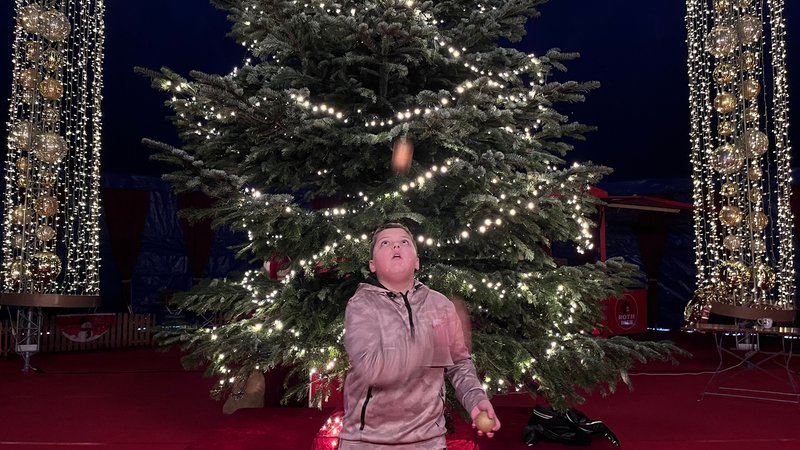 Ein Junge jongliert mit drei Bällen vor einem Weihnachtsbaum mit erleuchteter Lichterkette