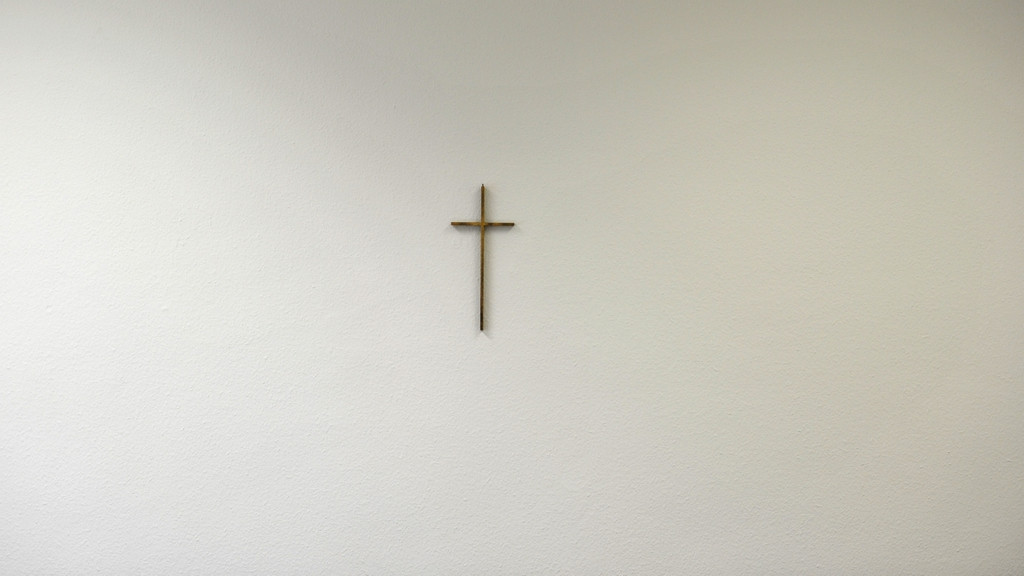 Ein schlichtes Metall-Kreuz auf einer ansonsten kahlen weißen Wand (Symbolbild)