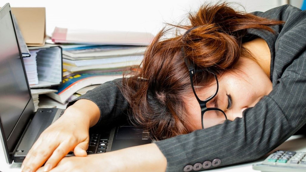 Eine Frau liegt erschöpft vor ihrem Laptop auf dem Schreibtisch (Symbolbild).