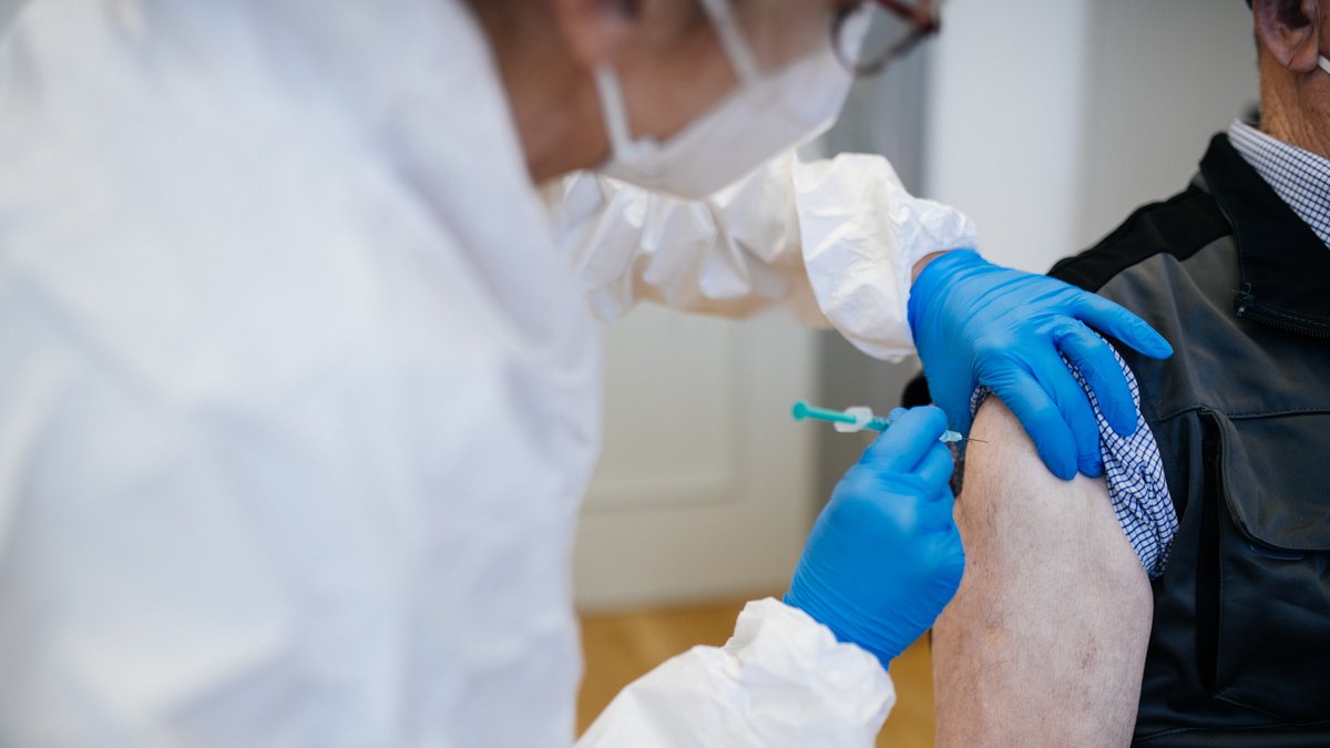 Ein medizinischer Mitarbeiter in weißer Schutzkleidung impft einen Patienten in den Oberarm.