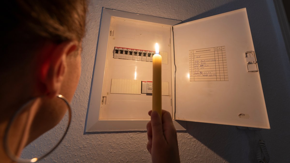 Eine Frau hält bei einem Stromausfall eine Kerze in der Hand und blickt in einen Sicherungskasten.
