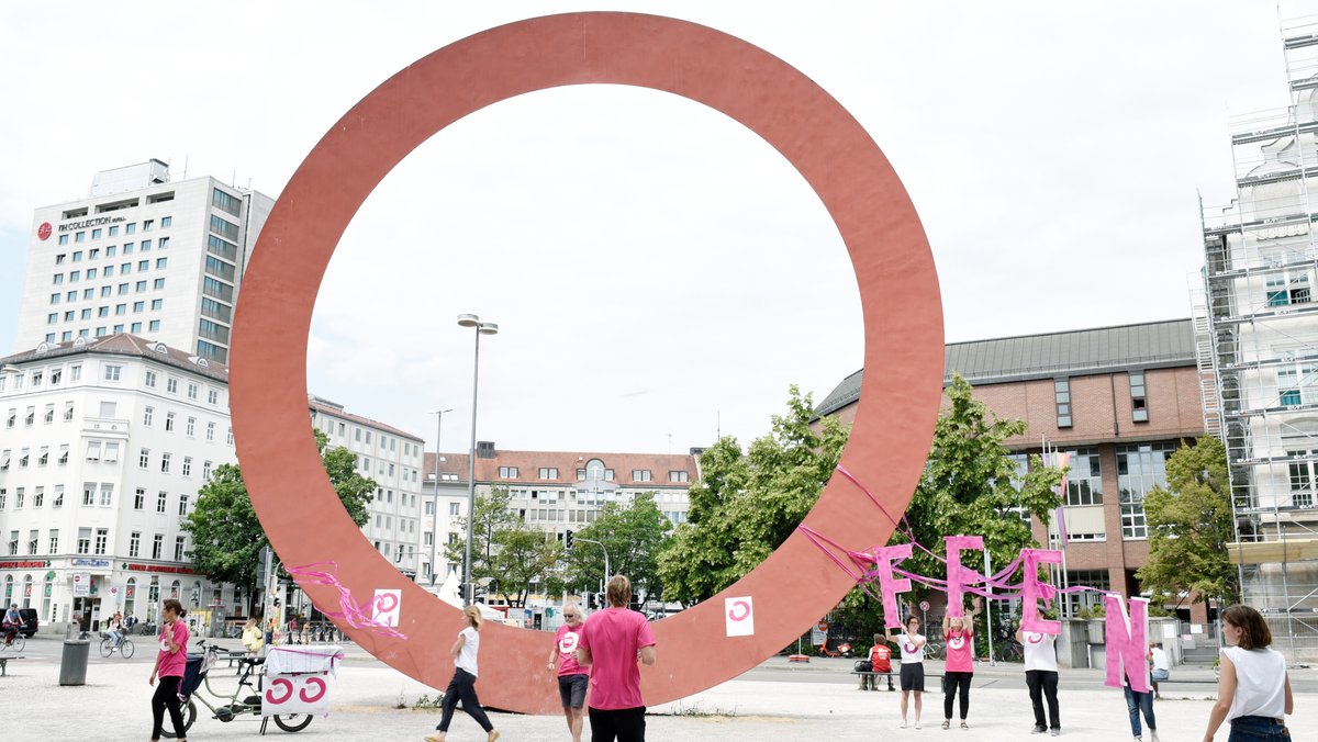 Die zwölf Meter hohe ringförmige Skulptur am Karl-Stützel-Platz am Alten Botanischen Garten in München, daneben Aktivisten von "Offen bleiben".