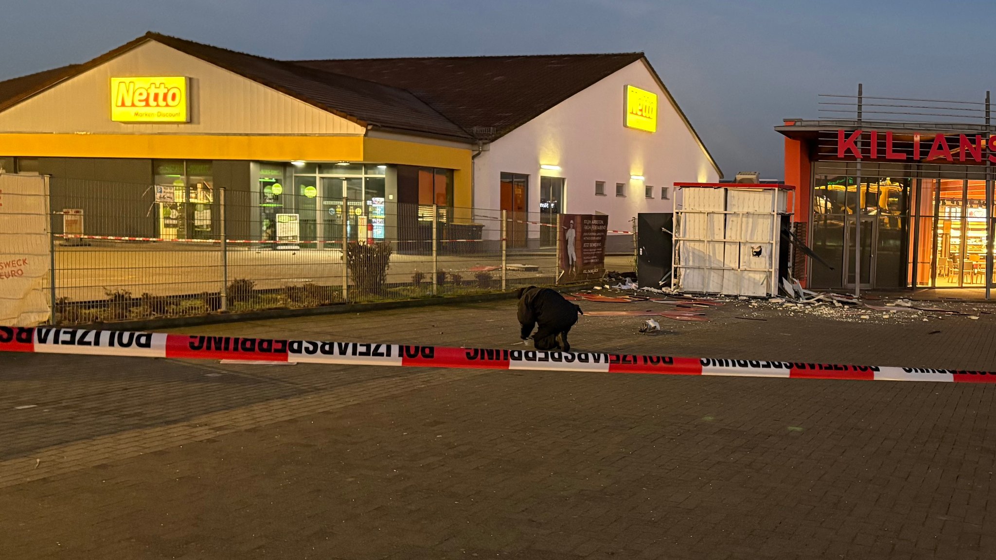 Flatterband sperrt den Tatort ab, dahinter ist ein Polizist bei der Spurensicherung. Im Bildhintergrund ist die Bäckereifiliale zu sehen und der Container der Sparkasse. Davor liegen die Reste des Geldautomaten auf dem Boden. | Bild: News5 Flatterband sperrt den Tatort ab, dahinter ist ein Polizist bei der Spurensicherung. Im Bildhintergrund ist die Bäckereifiliale zu sehen und der Container der Sparkasse. Davor liegen die Reste des Geldautomaten auf dem Boden.