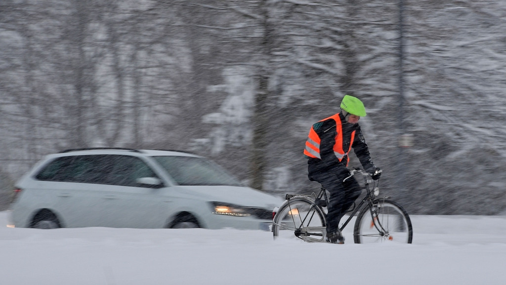 Ein Radler fährt bei Schneetreiben auf dem Radweg, daneben auf der Straße fährt ein Auto.