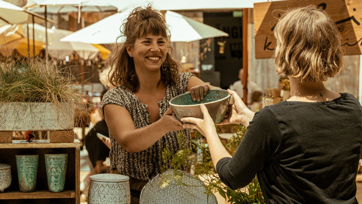 Junge Frau übergibt anderer Frau an einem Markstand eine Keramikschüssel 