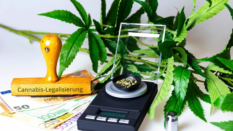 Eine Cannabis-Planze, davor eine Waage, Geldscheine und ein Stempel mit der Aufschrift "Cannabis-Legalisierung"