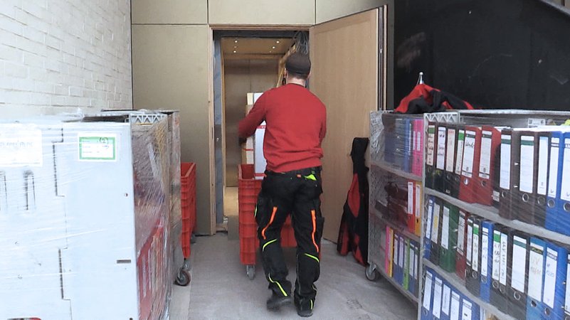 Ein Möbelpacker fährt einen Rollcontainer vorbei an Rollregalen mit Aktenordnern.