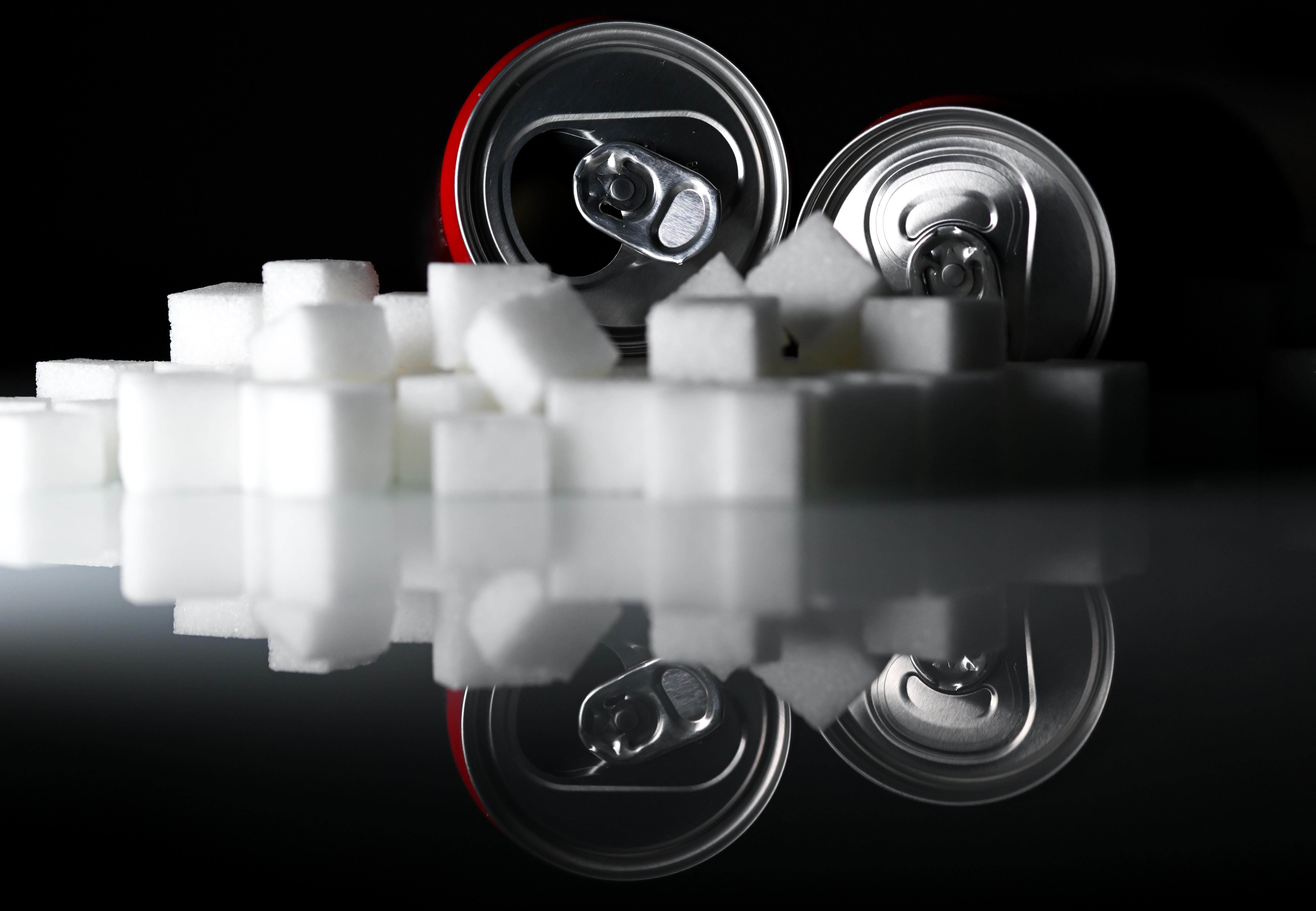 Zwei Getränkedosen mit Softdrinks und zahlreiche Zuckerwürfel liegen auf einem Tisch (Symbolbild).