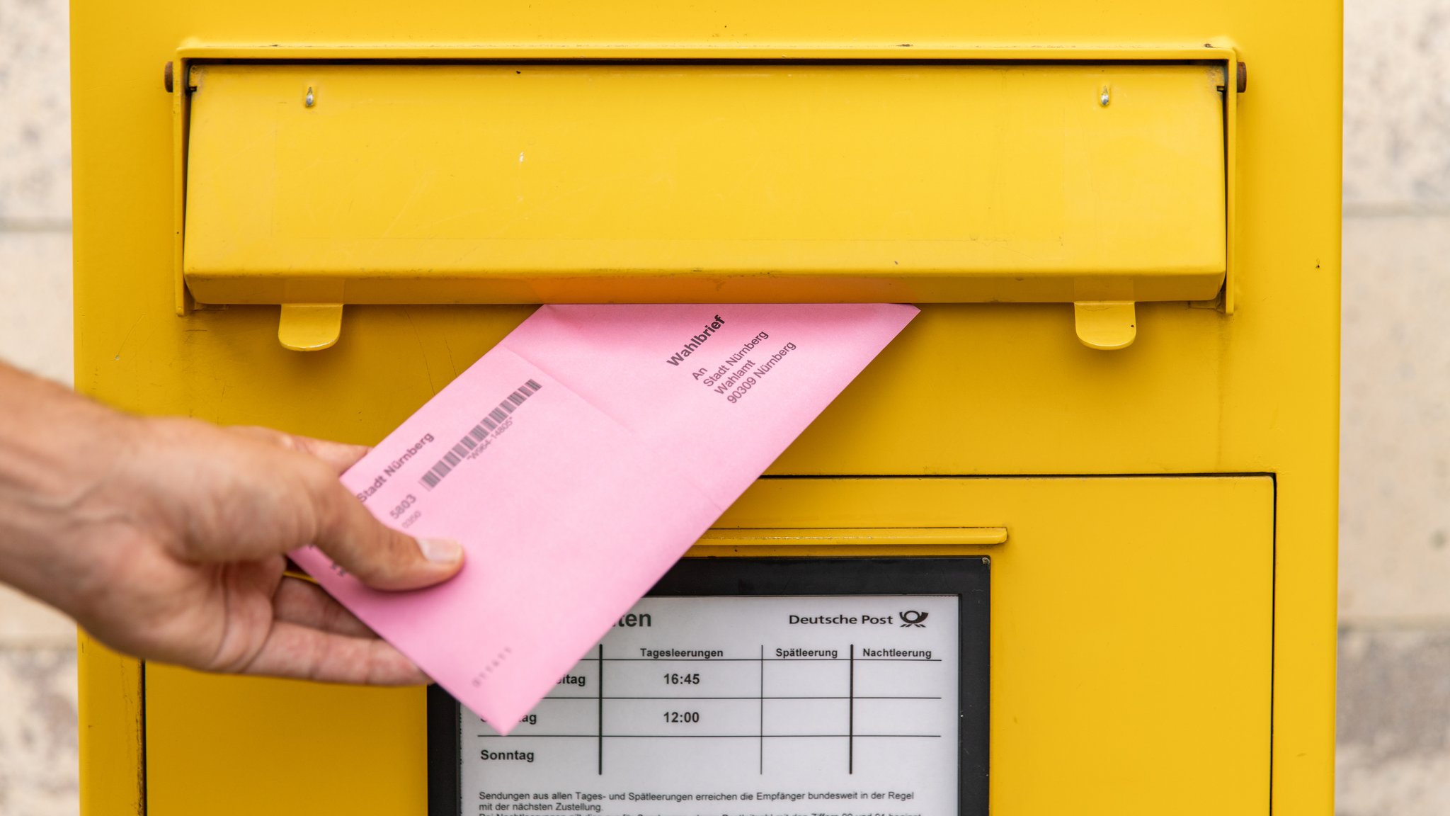 Zu sehen ist ein roter Wahlbrief, den eine Hand in einen gelben Briefkasten einwirft.