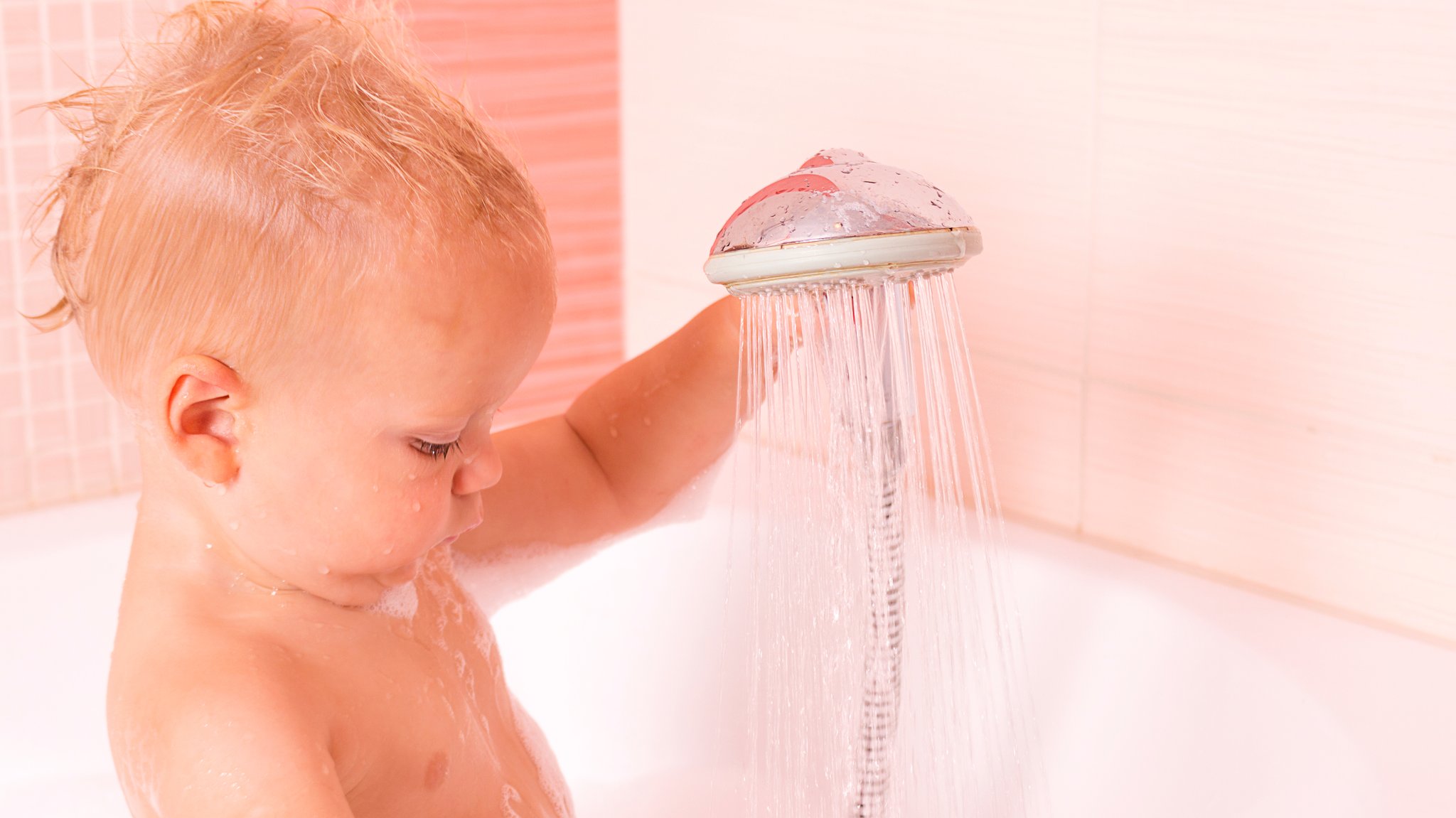 Blonder Babykopf im Profil mit einem laufenden Duschkopf.